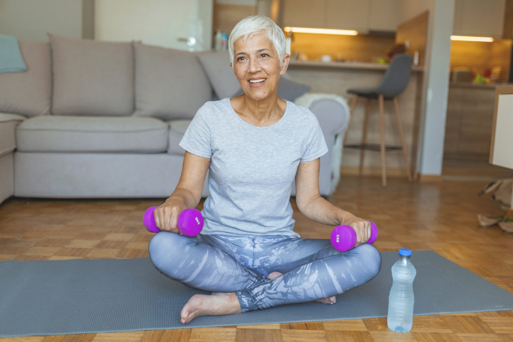 Portrait of senior woman sitting on exercise mat with dumbbells at home. Happy older woman in sports wear training with dumbbells indoors on the wi...