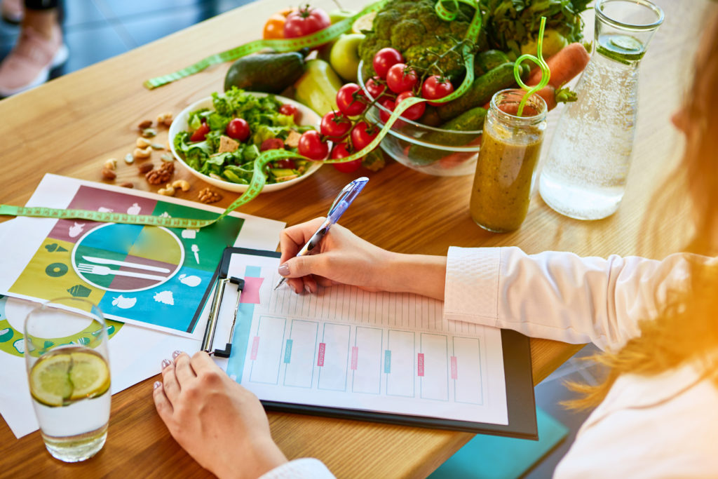 Woman dietitian in medical uniform with tape measure working on a diet plan sitting with different healthy food ingredients in the green office on ...