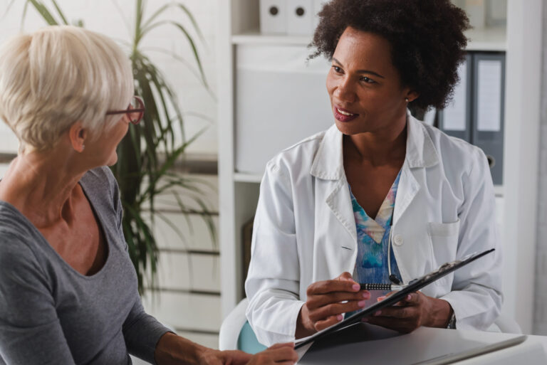 A female doctor sits at her desk and chats to an elderly female